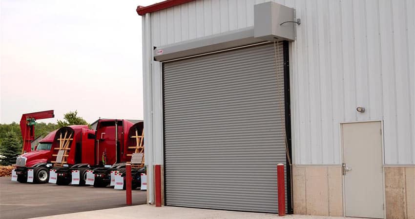Picture of a white warehouse with a silver garage door. Red trucks are in the background on the left.
